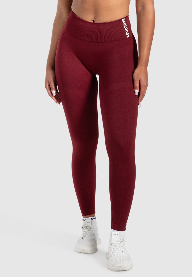 Leggings burdeos con una cintura alta, textura suave y un sutil logotipo en el lateral. Combinados con zapatillas deportivas blancas.