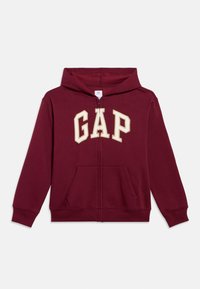 Tamnosmeđa dukserica na patent s mekanom teksturom. Ima kremasti logotip "GAP" na prsima, kangur džepove i standardni dizajn dukserice.