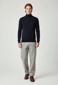 Navy Pullover mit hohem Kragen aus weichem Stoff, kombiniert mit hellgrauen Hosen und braunen Schnürschuhen. Einfaches Design und figurbetonte Form.
