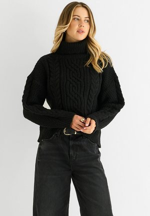 Black knitted sweater with a high turtleneck, featuring intricate cable patterns and wide sleeves, paired with dark denim pants.
