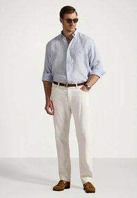 Homem a vestir uma camisa azul clara de botões, calças brancas, cinto castanho, sapatos loafers castanhos, óculos de sol e relógio de pulso, de pé com uma mão no bolso.