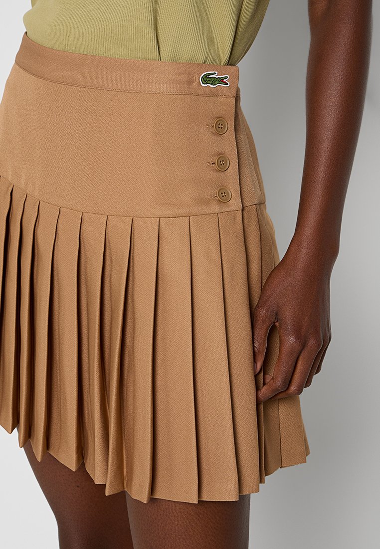 Gonna plissettata in tessuto beige con una texture liscia, dotata di una vita con bottoni e una patch con il marchio. Adatta per diverse occasioni.