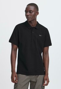 Junger Mann trägt ein schwarzes Poloshirt mit kurzen Ärmeln und "Blend"-Logo, kombiniert mit grauen Cargo-Hosen, steht vor einem schlichten Hintergrund.