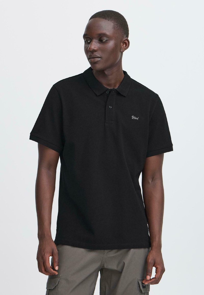 Junger Mann trägt ein schwarzes Poloshirt mit kurzen Ärmeln und "Blend"-Logo, kombiniert mit grauen Cargo-Hosen, steht vor einem schlichten Hintergrund.