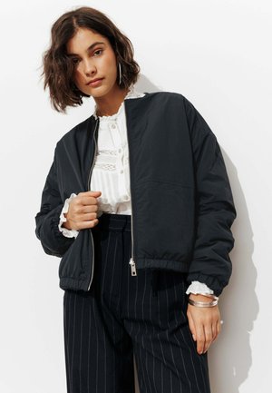 Femme aux cheveux courts et bouclés portant une veste zippée noire par-dessus un chemisier blanc et un pantalon noir à fines rayures, debout devant un fond blanc.