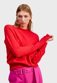 Pull en tricot rouge avec col rond, orné de petites clous décoratifs. Coupe ample et poignets côtelés. Associé à un pantalon rose vif et une ceinture.