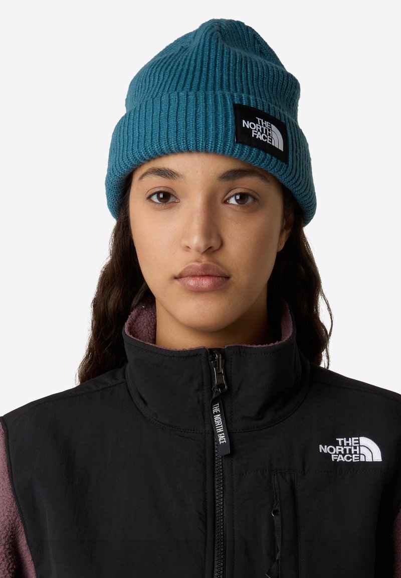 Teal strikket hue fra The North Face med ribbet tekstur og et sort logo-patch. Båret med en sort jakke med lynlås foran.