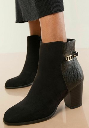 Schwarze Ankle Boots aus Wildleder mit einer strukturierten Reptilmuster-Optik, hohem Absatz und einem goldenen Akzent am Knöchel.