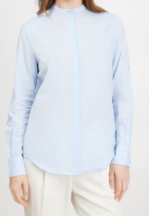 Jasnoniebieska bluzka z długim rękawem, z kołnierzykiem mandaryńskim i ukrytym zapięciem na guziki, noszona z kremowymi spodniami na stojącej osobie.