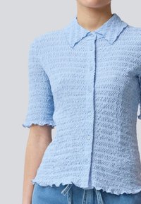Camisa de botão azul claro texturizada com um padrão ondulado, mangas curtas e uma bainha em forma de onda. Apresenta um colarinho pontudo e pequenos botões.