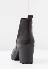 Zwarte leren enkellaars met een textuurfinish, ronde neus, hoge blokhak en elastische zijkanten. Achterlus voor eenvoudig aantrekken.