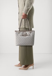 Sac à main gris à motifs avec deux anses supérieures, comportant des éléments en or et un détail de fermoir. Porté avec un pantalon couleur olive et des talons beiges.