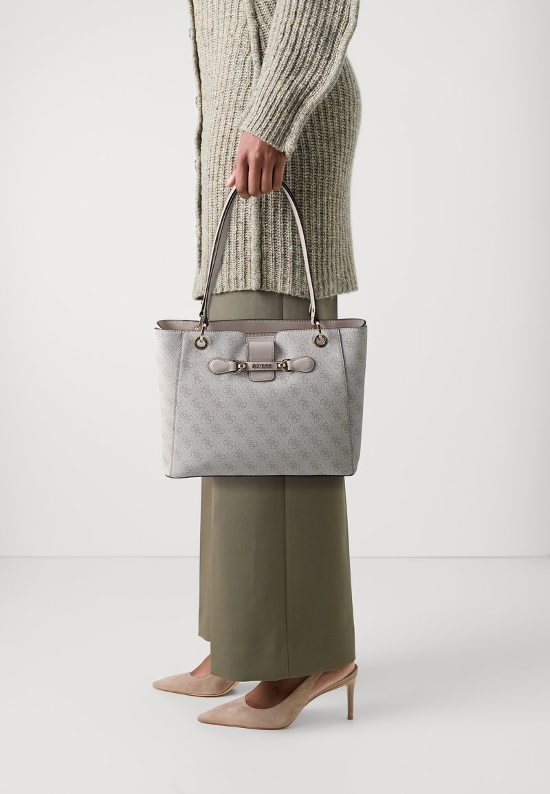 Sac à main gris à motifs avec deux anses supérieures, comportant des éléments en or et un détail de fermoir. Porté avec un pantalon couleur olive et des talons beiges.