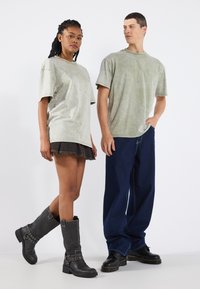 Dva modelové stojí vedle sebe. Žena má na sobě světle zelené oversized tričko, černou džínsovou sukni a šedé vysoké boty s pásky. Muž má na sobě odpovídající světle zelené oversized tričko a volné tmavě modré džíny.