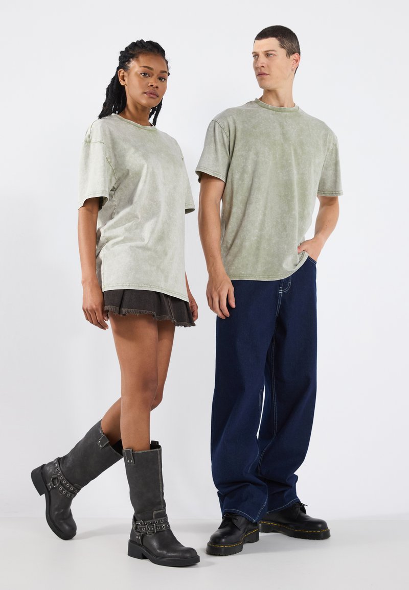 Dva modelové stojí vedle sebe. Žena má na sobě světle zelené oversized tričko, černou džínsovou sukni a šedé vysoké boty s pásky. Muž má na sobě odpovídající světle zelené oversized tričko a volné tmavě modré džíny.