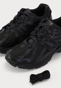 Paire de chaussures de course noires en mesh et cuir avec lacets noués, une paire supplémentaire de lacets noirs placée à côté sur une surface blanche.