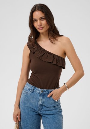 Femme portant un haut marron à une épaule avec des volants et un jean bleu, une main dans la poche et tenant un petit sac dans l'autre.