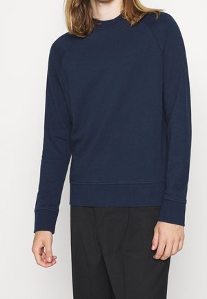 Sudadera navy con largas mangas raglán, cuello redondo y un acabado texturizado. Presenta un dobladillo y puños acanalados. Lleva puesta sobre pantalones negros.