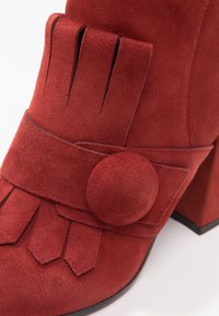 Chaussure en suède rouge avec talon bloc, ornée de franges et d'un grand bouton décoratif rond sur la bride avant.