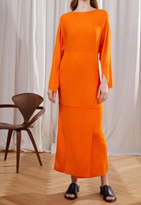 Orange långärmad klänning med en strukturerad silhuett och sidofickor, gjord av ett mjukt tyg; kombinerad med svarta slip-on skor.