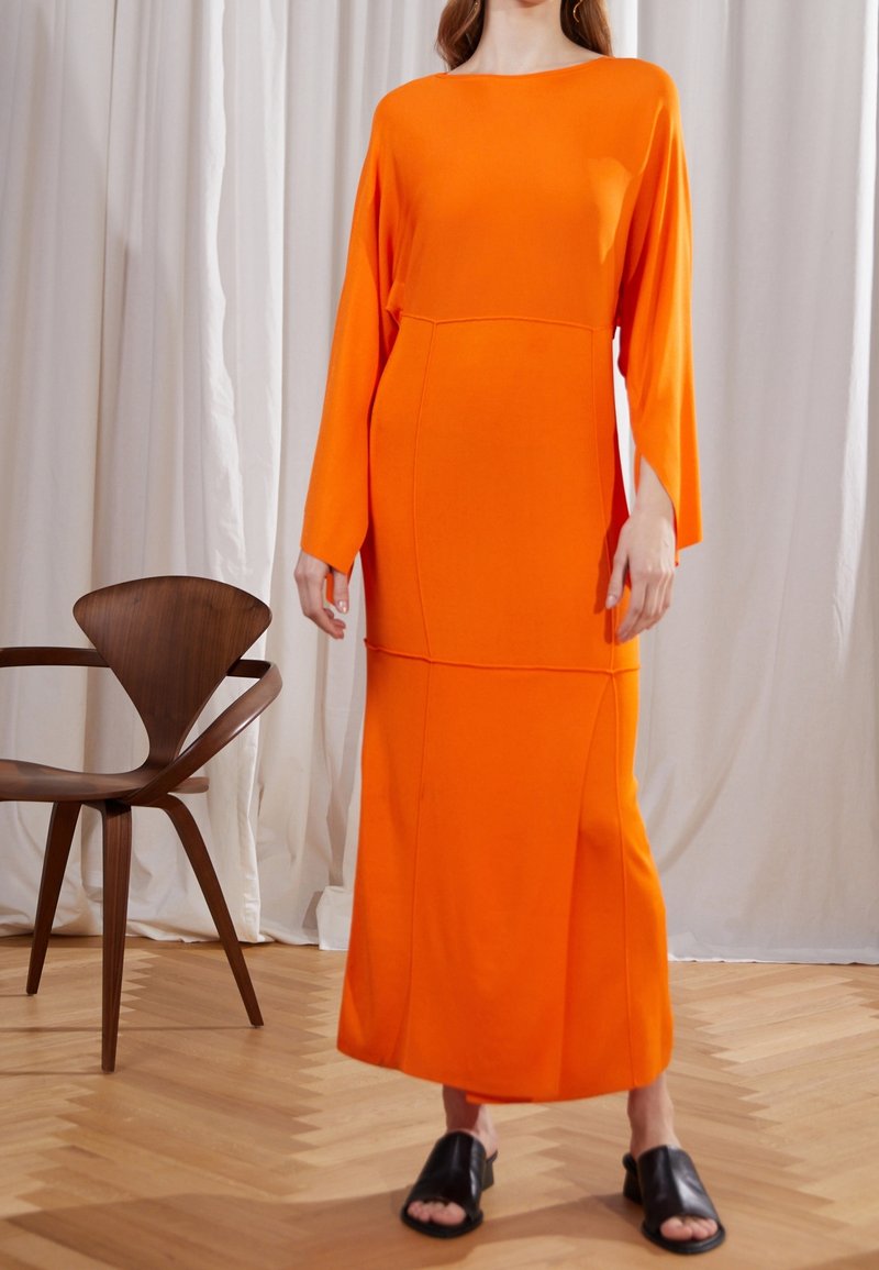 Orange långärmad klänning med en strukturerad silhuett och sidofickor, gjord av ett mjukt tyg; kombinerad med svarta slip-on skor.
