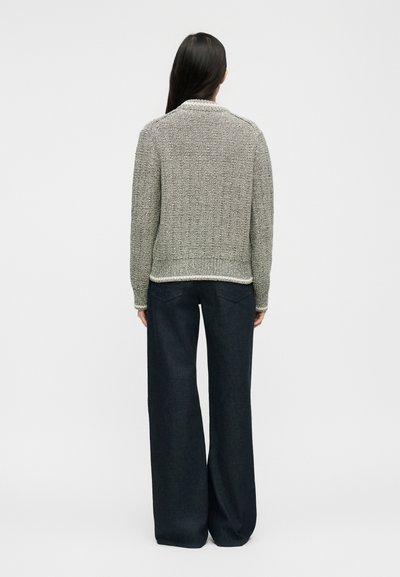 Γυναίκα με μακριά σκούρα μαλλιά που φοράει ανοιχτογκρι πλεκτό πουλόβερ και φαρδιά σκούρα παντελόνια, όρθια με την πλάτη γυρισμένη σε καθαρό λευκό φόντο.