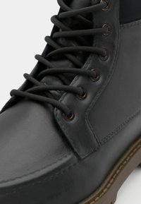 Bota de cuero negro con cordones redondos, ojales de metal y una superficie texturizada. La suela es marrón con un detalle de costura visible.