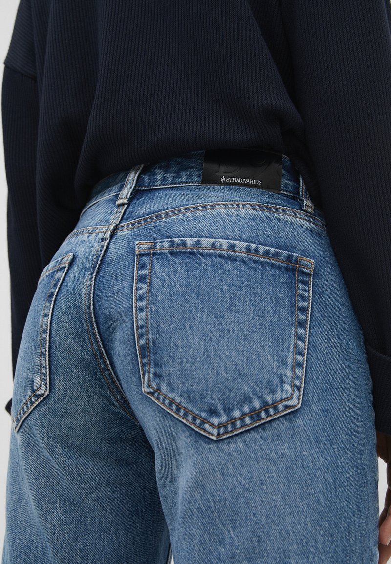 Vaqueros de denim azul con un corte clásico, diseño de cinco bolsillos, costuras visibles y una etiqueta de marca negra en la cinturilla.