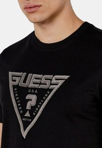 T-shirt in cotone nero con logo triangolare che presenta la scritta "GUESS" in lettere grasse, accompagnato da ricami grigi e un dettaglio a forma di punto interrogativo.