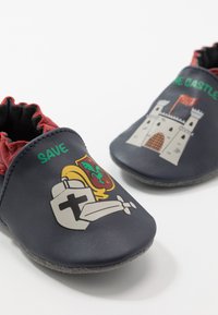 Bottines en cuir bleu marine avec des ouvertures en élastique rouge, présentant des graphismes de château et de chevalier, ainsi que le texte "SAUVEZ LE CHÂTEAU" en vert sur les côtés.