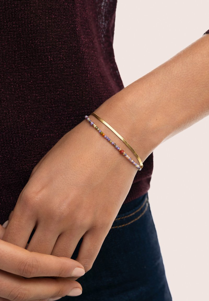 Goldfarbener Metall-Cuff-Armband neben einem Perlenarmband mit kleinen, mehrfarbigen Perlen, getragen an einer Hand vor einem dunklen Hemd.