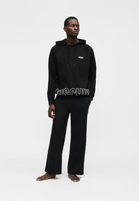 Mann stående barbeint iført svart Moschino-hettegenser med hvit logo og matchende svarte vide bukser mot en enkel lys bakgrunn.