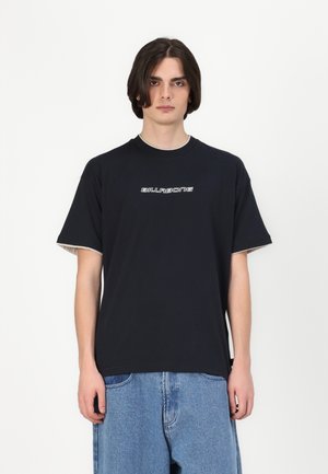 Czarny t-shirt z krótkim rękawem z białym logo "BILLABONG", okrągłym dekoltem i kontrastującym jasnoszarym wykończeniem przy rękawach. Noszony z jasnoniebieskimi dżinsami.