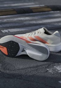 Λευκά και πορτοκαλί παπούτσια για τρέξιμο Adidas με ορατές σόλες Lightstrike που τοποθετούνται σε άσφαλτο κοντά σε διάβαση πεζών.