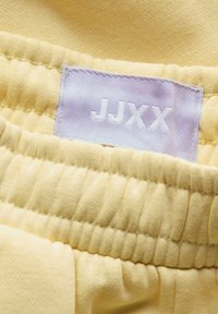 Gul stoff med en myk tekstur som har en samlet midje. En lilla vevd etikett med "JJXX" i hvit er synlig.