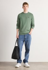 Green long-sleeve sweater with round neck and black logo, paired with blue jeans and white sneakers. Carrying a black tote bag.