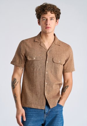 Giovane uomo con capelli ricci, indossa una camicia marrone a maniche corte con bottoni e jeans blu, in piedi con una mano in tasca.