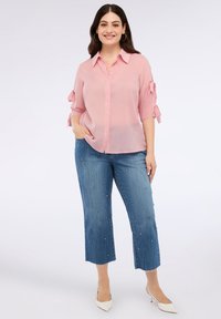 Jasnoróżowa koszula z guzikami z podwiniętymi rękawami zawiązana na łokciach, zestawiona z niebieskimi, krótkimi dżinsami ozdobionymi kolorowymi detalami.
