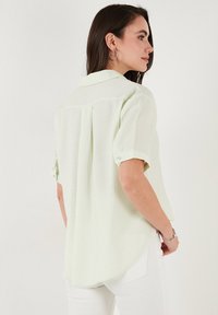 Blusa a maniche corte di un verde chiaro con una consistenza morbida, polsini a bottoni, piega sul retro e orlo curvo. Progettata per un fit rilassato.