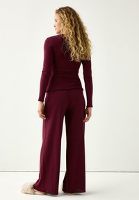 Burgundy ribbad stickad topp och vida byxor med fransade fållar; mjuk textur synlig. Modellen står med ryggen vänd, vilket visar upp plaggets design.