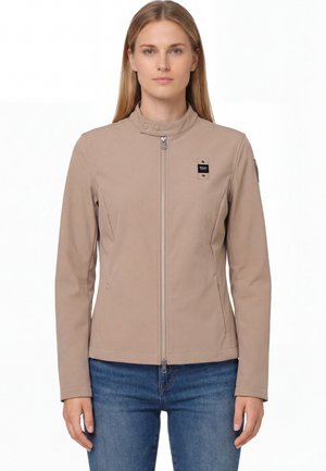 Kobieta ubrana w beżową kurtkę na zamek z wysokim kołnierzem i dwoma przednimi kieszeniami, w połączeniu z niebieskimi dżinsami, stojąca na białym tle.