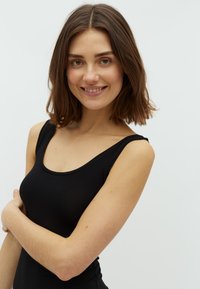 Melnā bezpiešu topiņa ar apaļu kakla izgriezumu, gluda tekstūra, pieguļoša forma, izceļ plecus un augšējo ķermeni. Vienkārša fona.