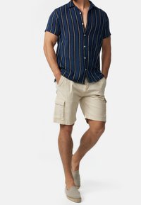 Man in een blauwe korte mouw met verticale strepen, beige cargoshorts met zakken en beige instap schoenen. Handen in de zakken.