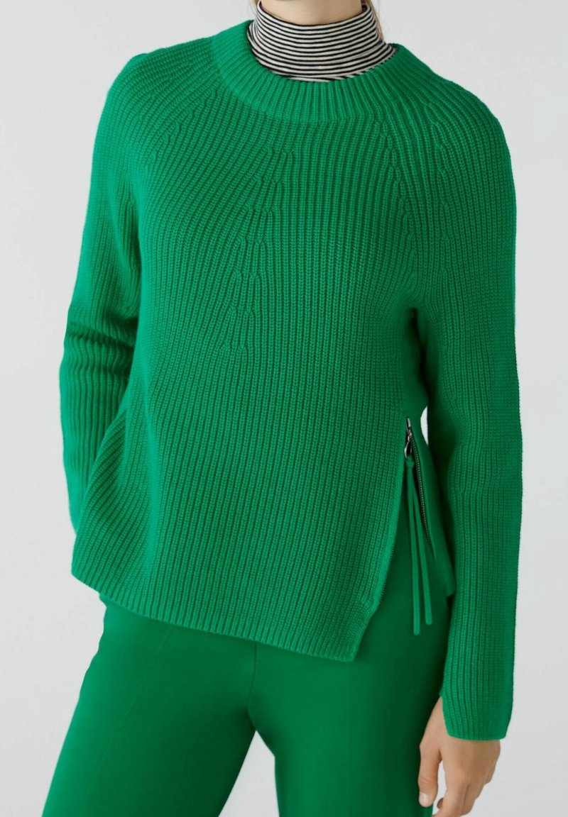 Pull en maille verte avec une texture côtelée, col rond et détail de fermeture éclair sur le côté. Stylé avec un col roulé rayé en dessous.
