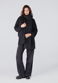 Zwarte oversized trui met een gestructureerde afwerking en subtiele glans, gecombineerd met wijde donkere jeans en robuuste zwarte laarzen.