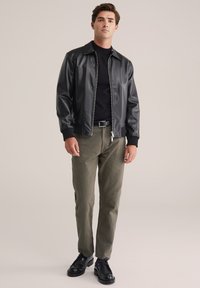 Joven de pie, vistiendo una chaqueta de cuero negra, un suéter negro, pantalones verde olivo, cinturón negro y zapatos negros, contra un fondo beige.