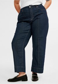 Tummansiniset denim-housut, joissa on rento istuvuus, korkea vyötärö, suora lahje ja kontrastiväriopastukset.