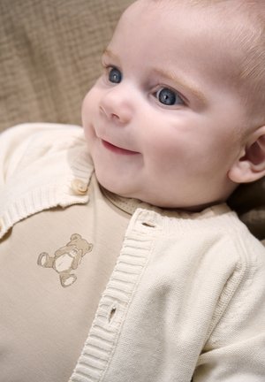 Licht crème vestje met geribbelde randen over een beige romper met een lichtbruine teddybeertafereel. Gladde stof, zachte textuur.