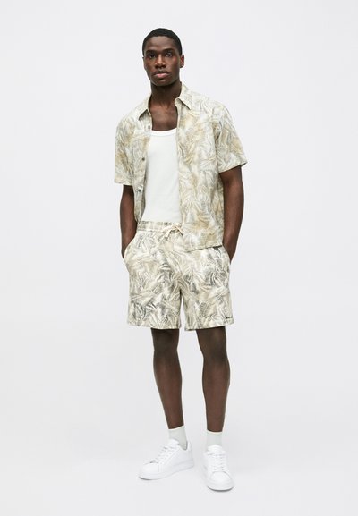 Chemise à manches courtes avec un motif de feuilles de palmier beige clair, associée à un short assorti et des baskets blanches. La chemise a une coupe décontractée ; mélange de coton.