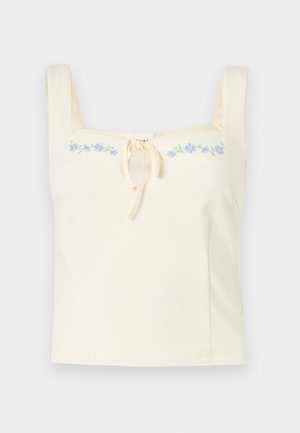 Κρεμ αμάνικη κοντή μπλούζα με τετράγωνο ντεκολτέ, λεπτομέρεια δέσιμου στο κέντρο και μπλε κεντητό λουλουδένιο διακοσμητικό στο στήθος.